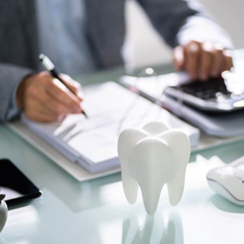 someone working with pen, paper, and calculator to determine cost at desk with computer keyboard, mouse, and large fake tooth